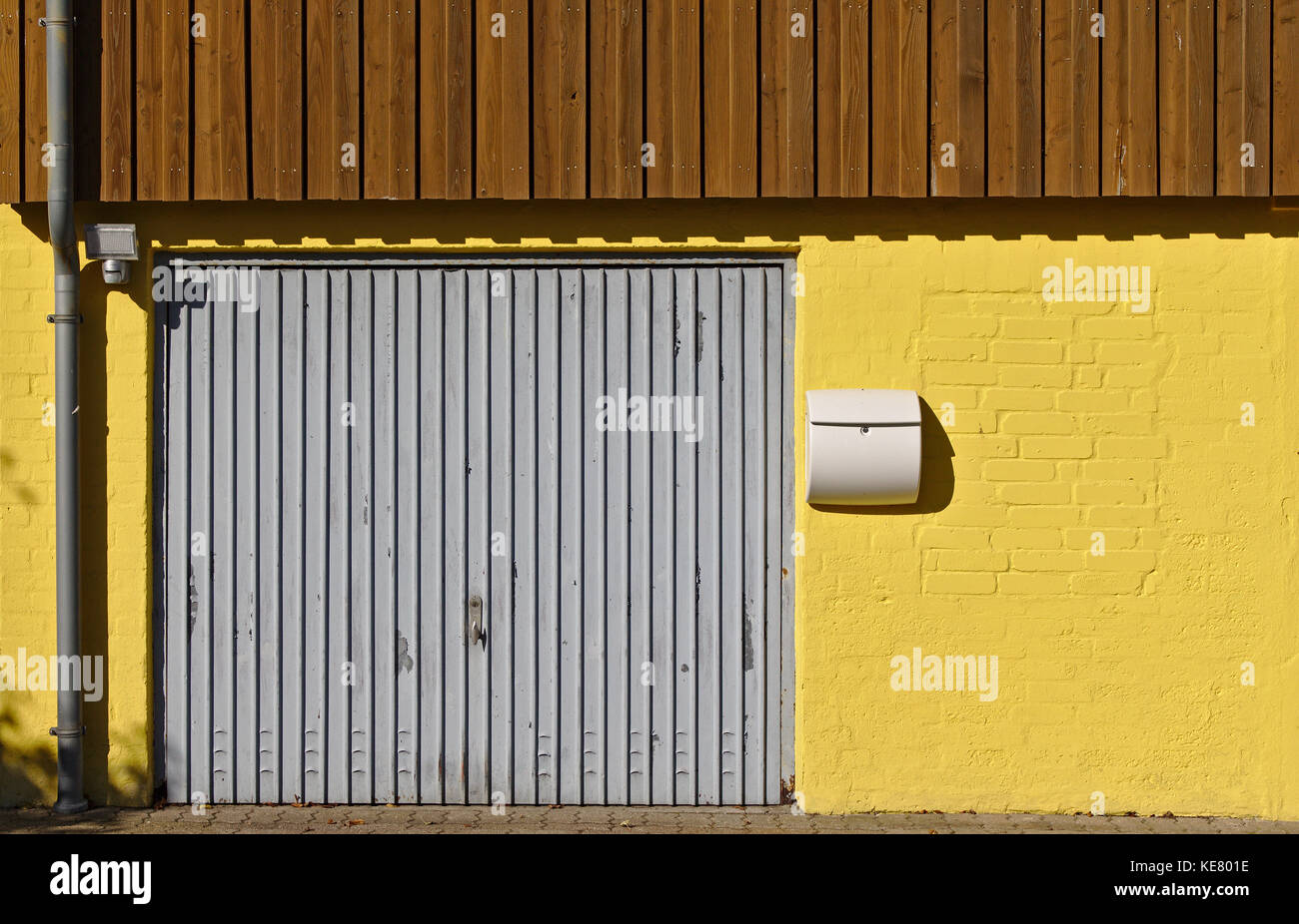Pared de ladrillo de color amarillo con la puerta del garaje, casilla de correo, tubo de bajante marrón y paneles de madera en el segundo piso Foto de stock