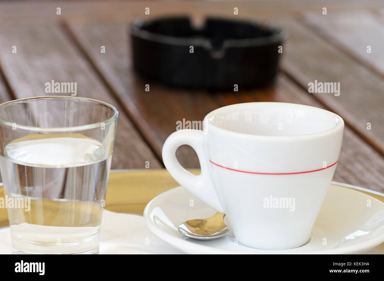 La foto de una taza de café en un plato con una cuchara junto a un vaso de agua y un cenicero con un fondo borroso Foto de stock