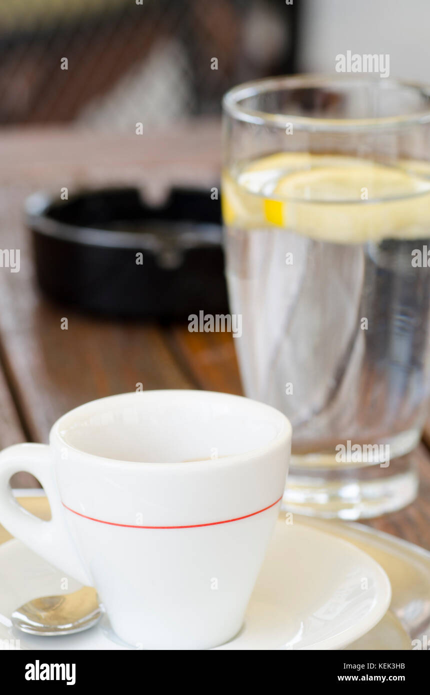 La foto de una taza de café en un plato con una cuchara junto a un vaso de agua y un cenicero con un fondo borroso Foto de stock