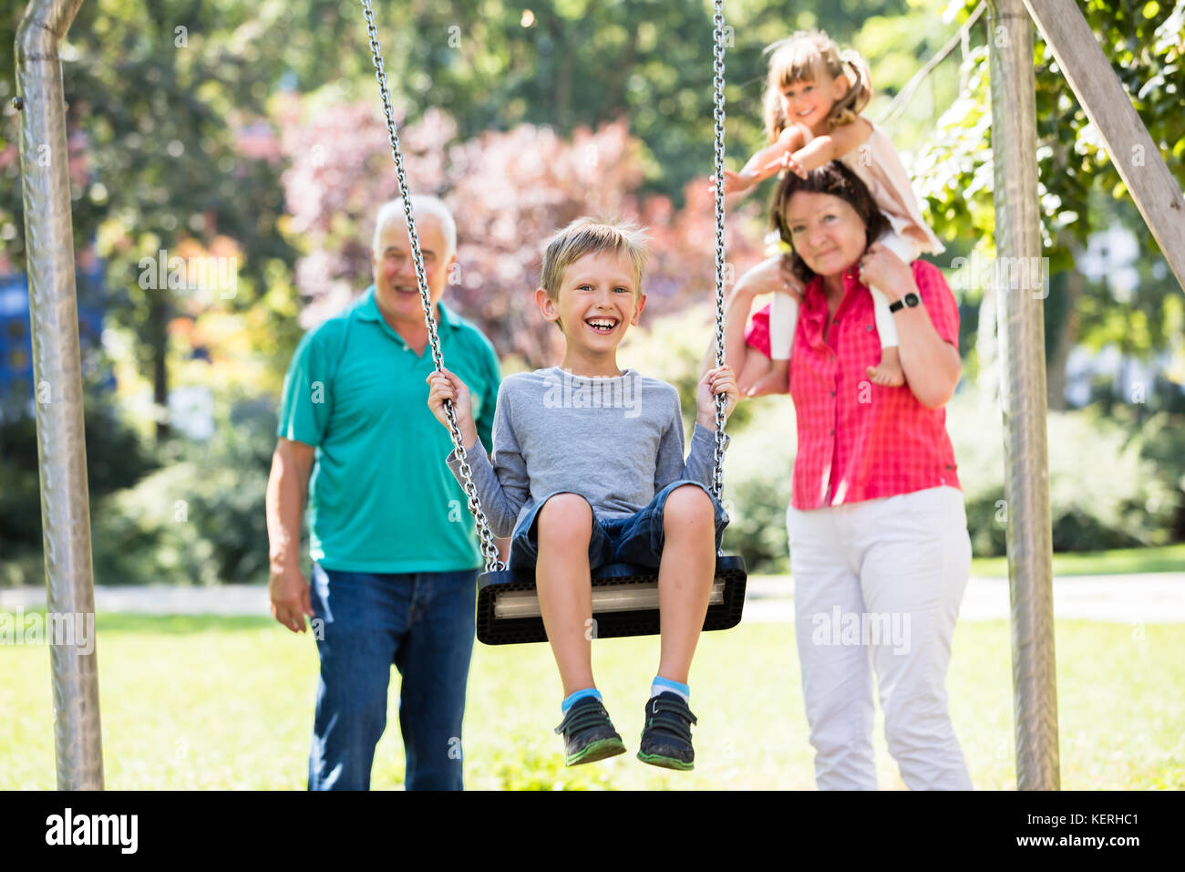 Feliz nieto kid divirtiéndose en swing con viejos abuelos de pie detrás en el parque Foto de stock