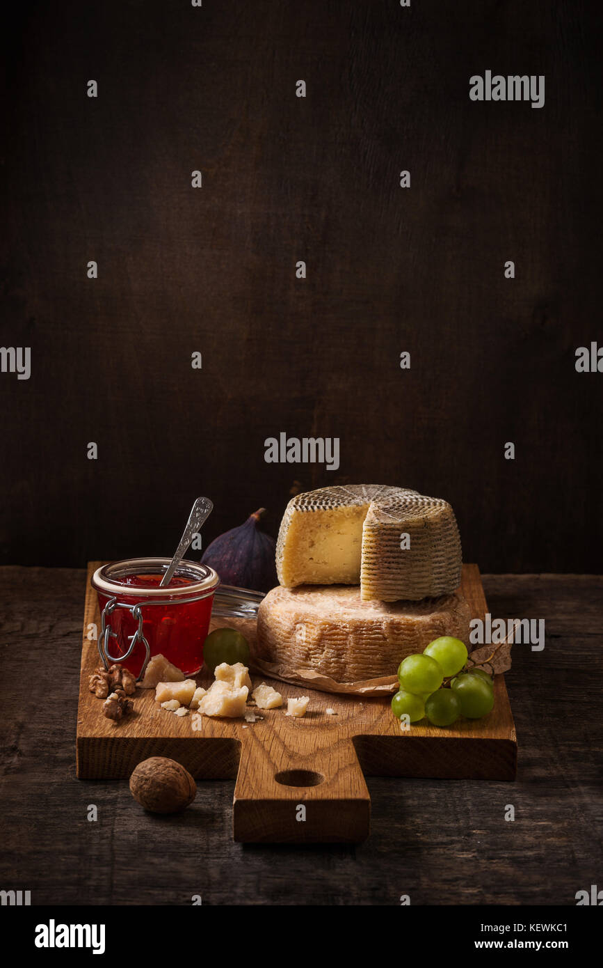 Dark bodegón con una tabla de quesos, fruta y vino en el fondo de madera áspera Foto de stock
