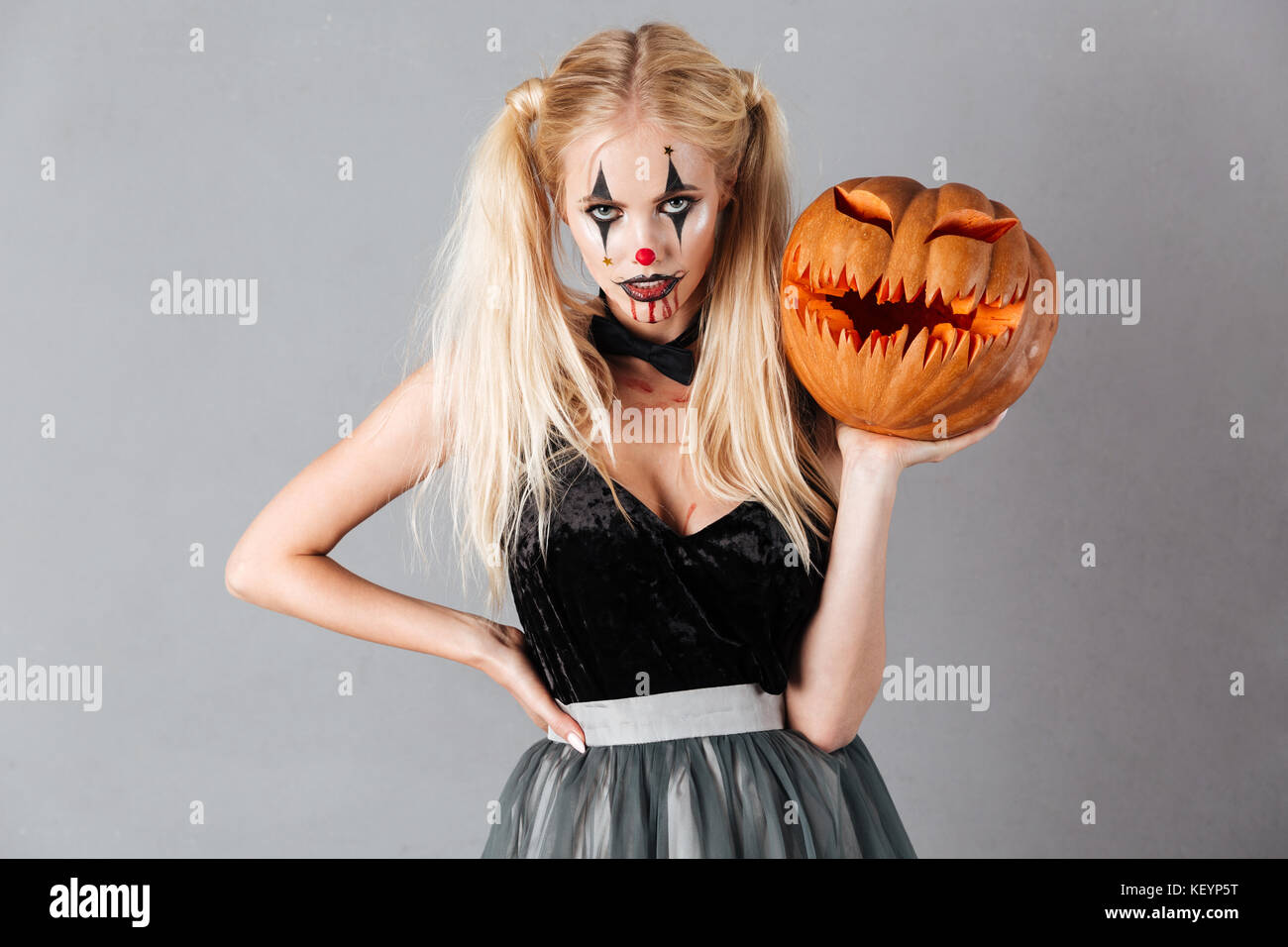 Atractiva mujer rubia en halloween maquillaje de payaso y vetas de sangre  mantiene curvado de calabaza y mirando a la cámara aislada sobre fondo gris  Fotografía de stock - Alamy