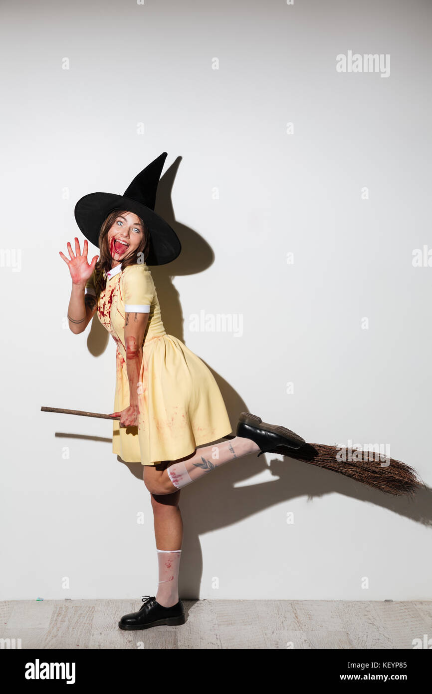 Longitud completa imagen de mujer feliz en disfraz de Halloween con la boca  abierta como la Bruja volando en escoba y mirando la cámara sobre fondo  blanco Fotografía de stock - Alamy