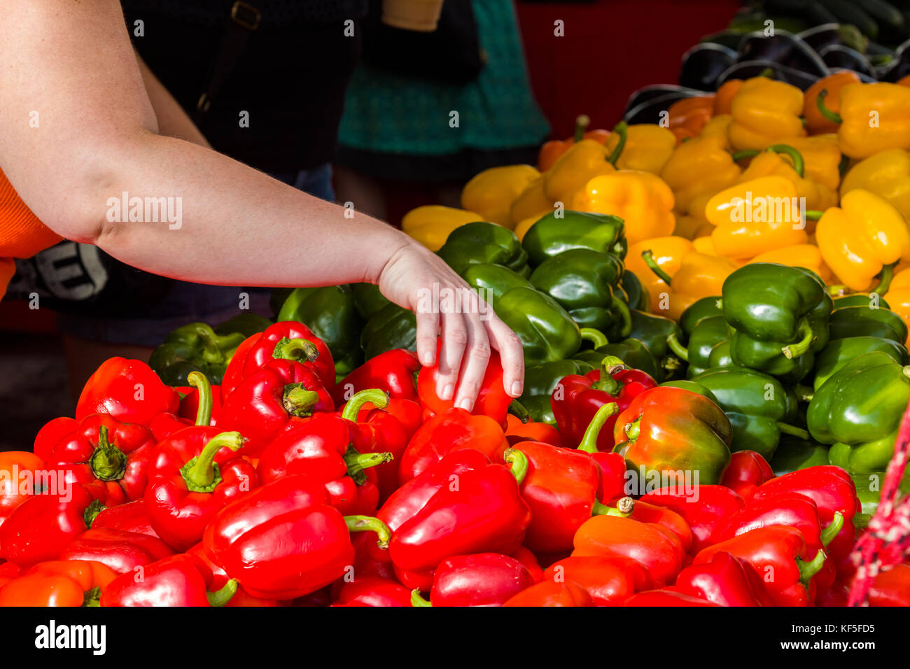 Mano alcanzando para una pimienta en un montón de pimientos rojos, verdes y amarillos en un mercado Foto de stock