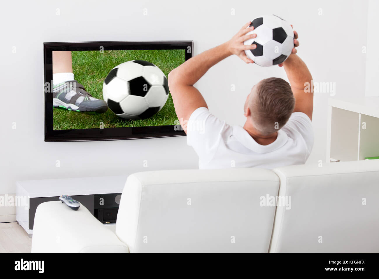 Emocionados hinchas de fútbol viendo un por televisión sosteniendo un balón de fútbol por de su como él está sentado en un cómodo sofá en su sala de estar