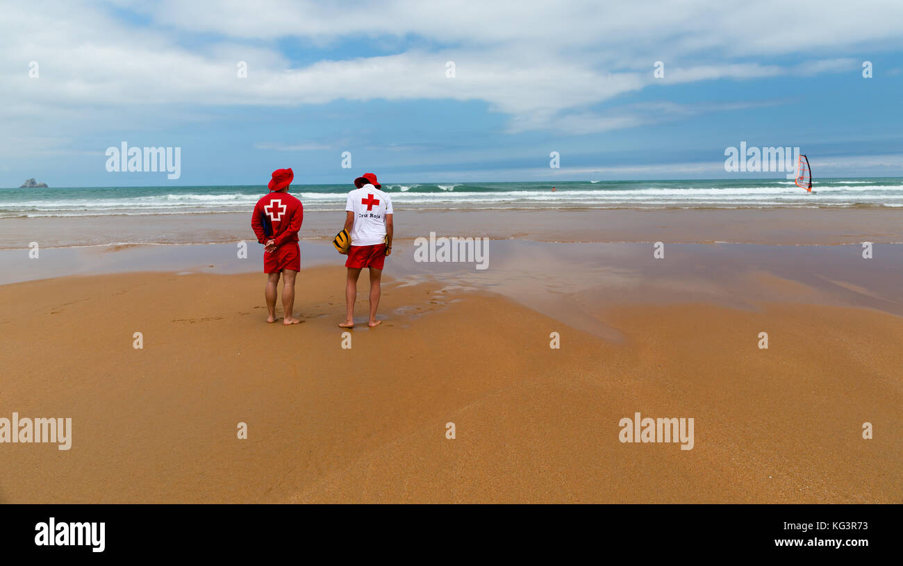 Orinon, España - Julio 6, 2016: dos socorristas desconocidos en la desierta playa. Los socorristas en rojo-blanco ropa colócate frente al océano en las olas de la EFS. Foto de stock