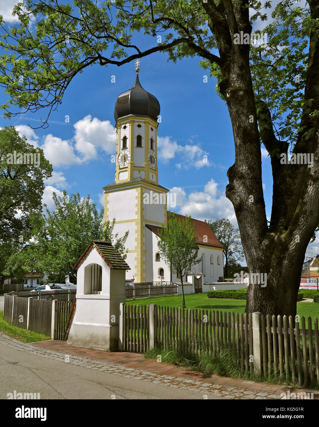 Alemania, Baviera, Aying, iglesia del pueblo, aguja bulbosa, calle, árbol, cerca, cielo blanco/azul, primavera Foto de stock