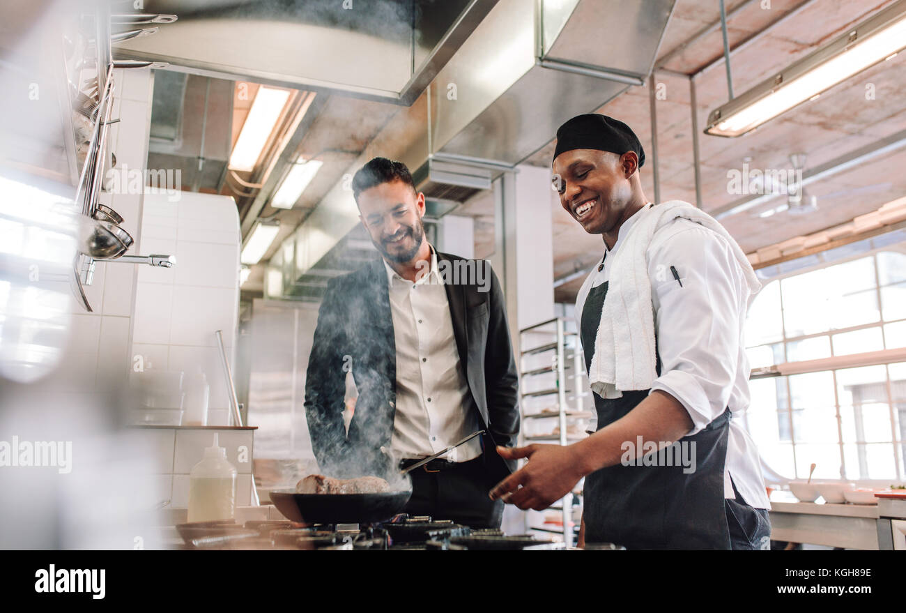 Sonriente chef cocinando comida sobre la estufa con el gerente del restaurante en pie. Gerente de restaurante con cocina en la cocina comercial. Foto de stock