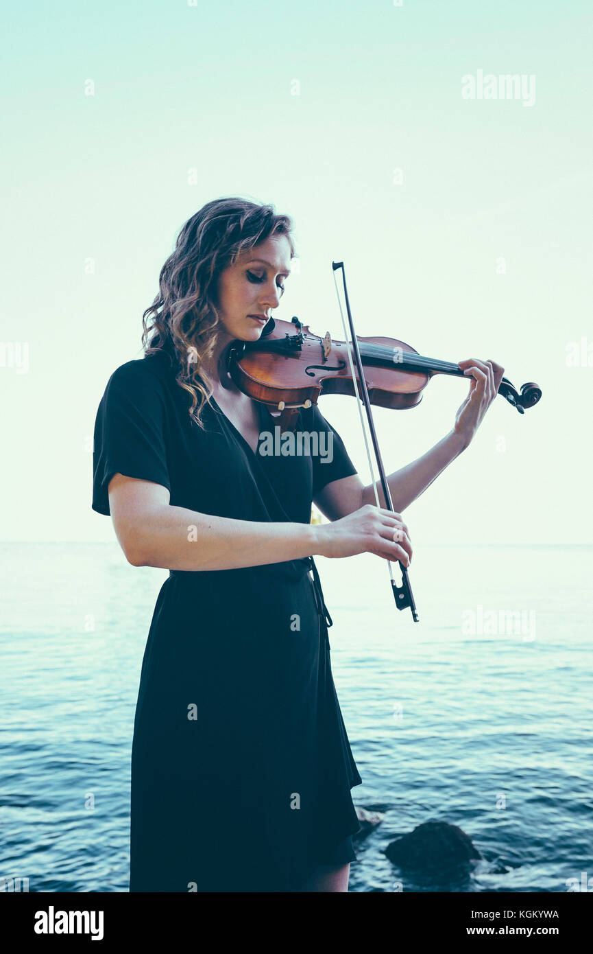 Hermosa joven a tocar el violín mientras está de pie por el lago contra el cielo claro Foto de stock