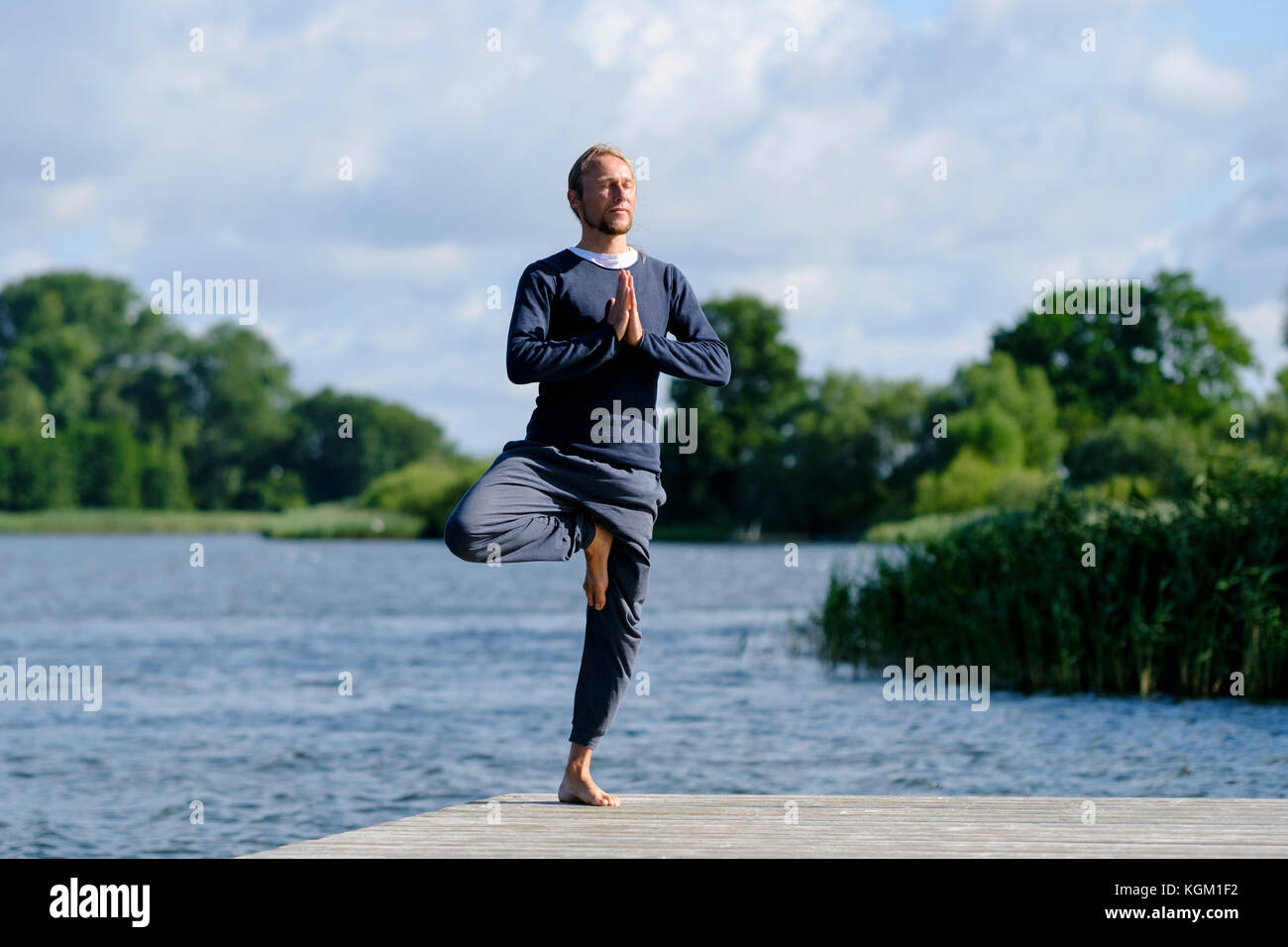 Hombre maduro, practicando la postura del árbol yoga en el muelle por el lago contra el cielo Foto de stock