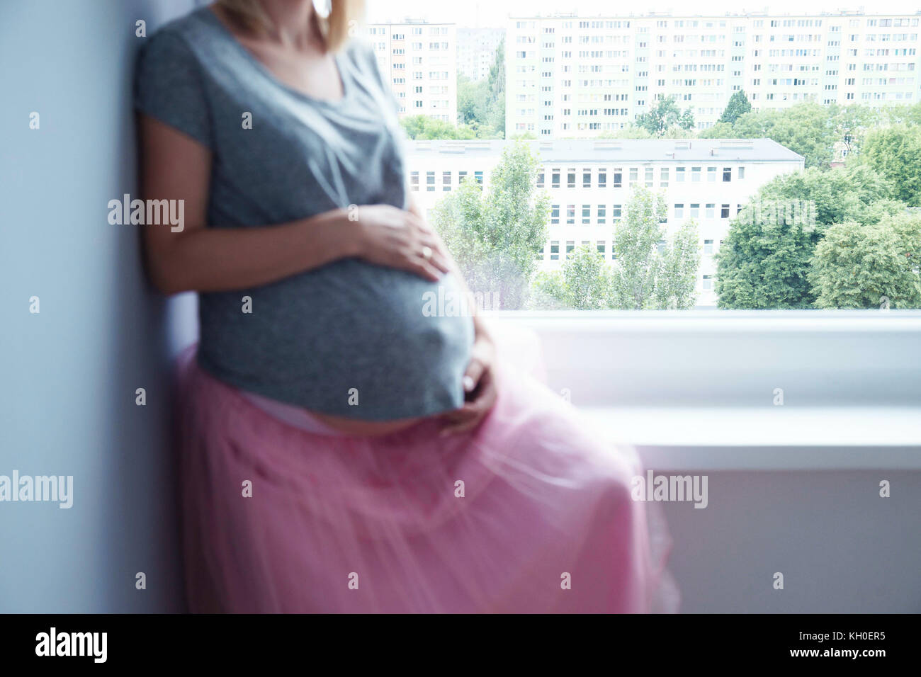 Anonymous mujer embarazada sentada sobre un alféizar de una falda rosa y una camiseta, centrarse en el fondo. Foto de stock