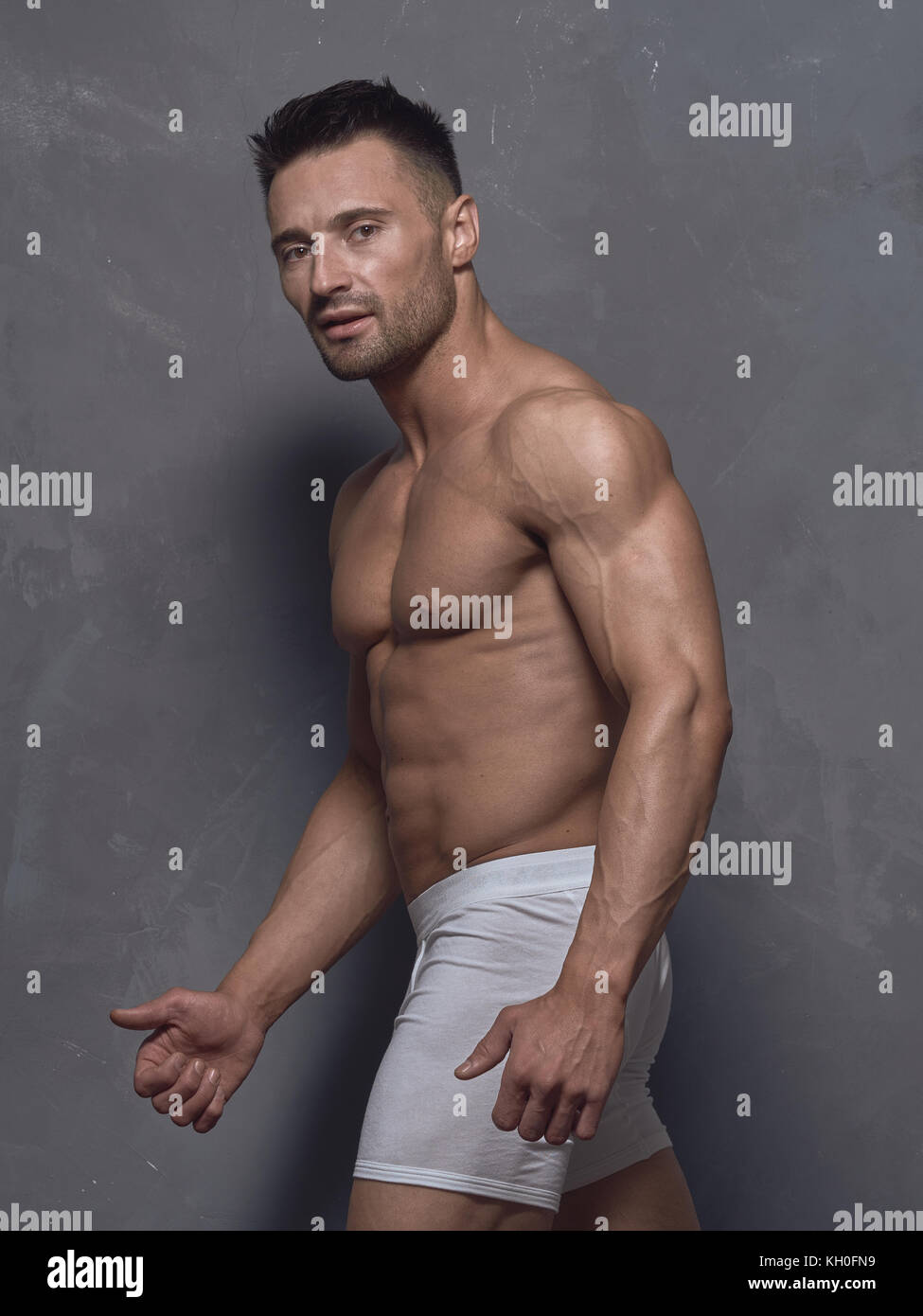 Un hombre de pelo negro con un corte de pelo corto está parado cerca de la  pared texturizada gris. El tipo tiene fuertes brazos y hombros musculares.  Su cuerpo está desnudo Fotografía