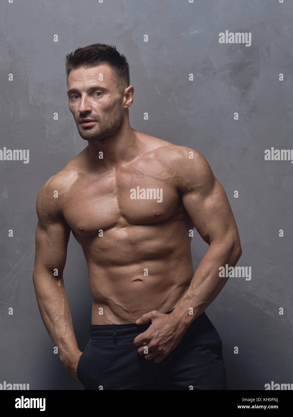 Un hombre de pelo negro con un corte de pelo corto está parado cerca de la  pared texturizada gris. El tipo tiene fuertes brazos y hombros musculares.  Su cuerpo está desnudo Fotografía