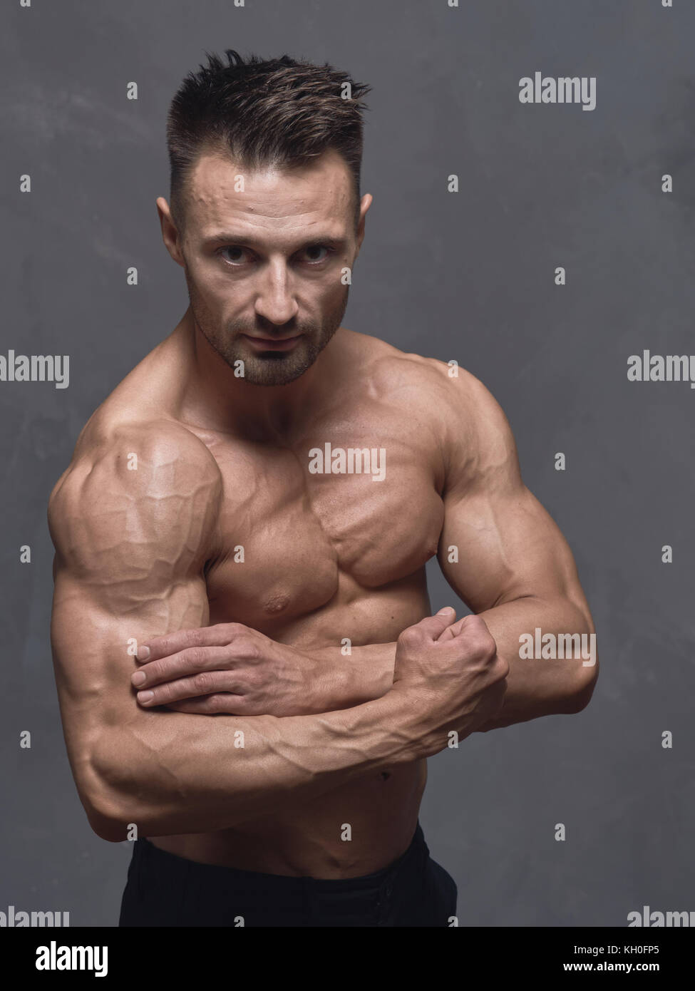 Un Hombre De Pelo Negro Con Un Corte De Pelo Corto Esta De Pie Cerca De La Pared Con Textura Gris El Chico Tiene Fuertes Musculosos Brazos Y Hombros Su Cuerpo Esta