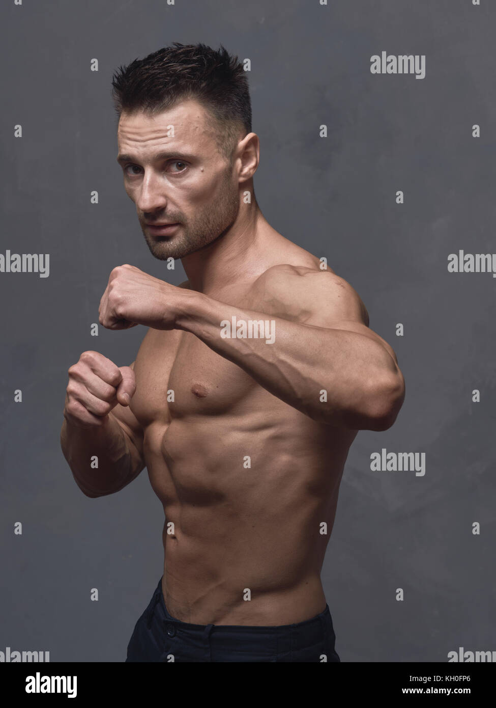 Un hombre de pelo negro con un corte de pelo corto está parado cerca de la  pared texturizada gris. El tipo tiene fuertes brazos y hombros musculares.  Su cuerpo está desnudo Fotografía