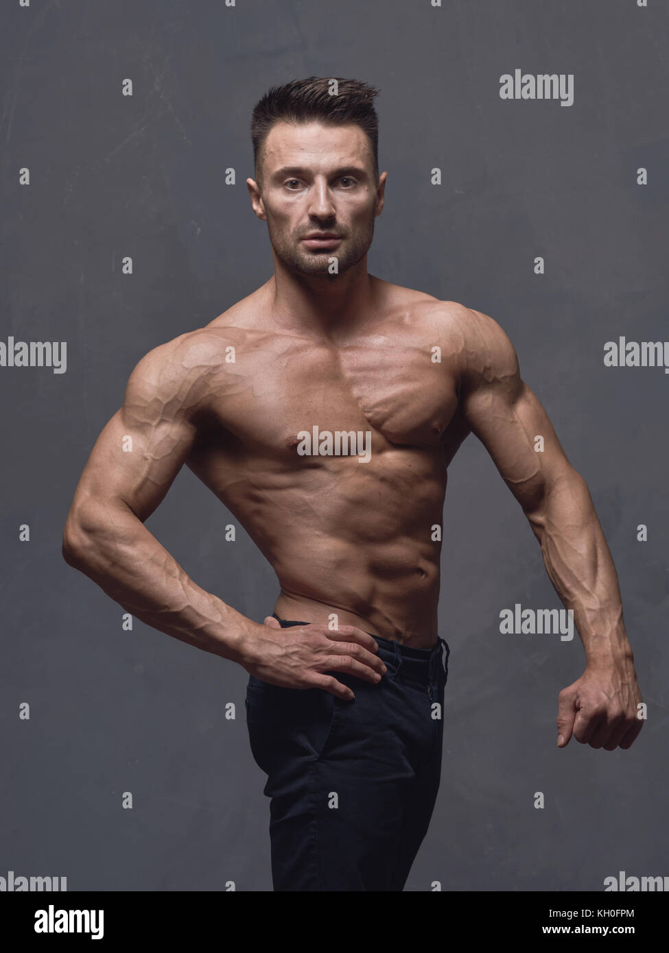 Un hombre de pelo negro con un corte de pelo corto está parado cerca de la  pared texturizada gris. El tipo tiene fuertes brazos y hombros musculares.  Su cuerpo está desnudo Fotografía