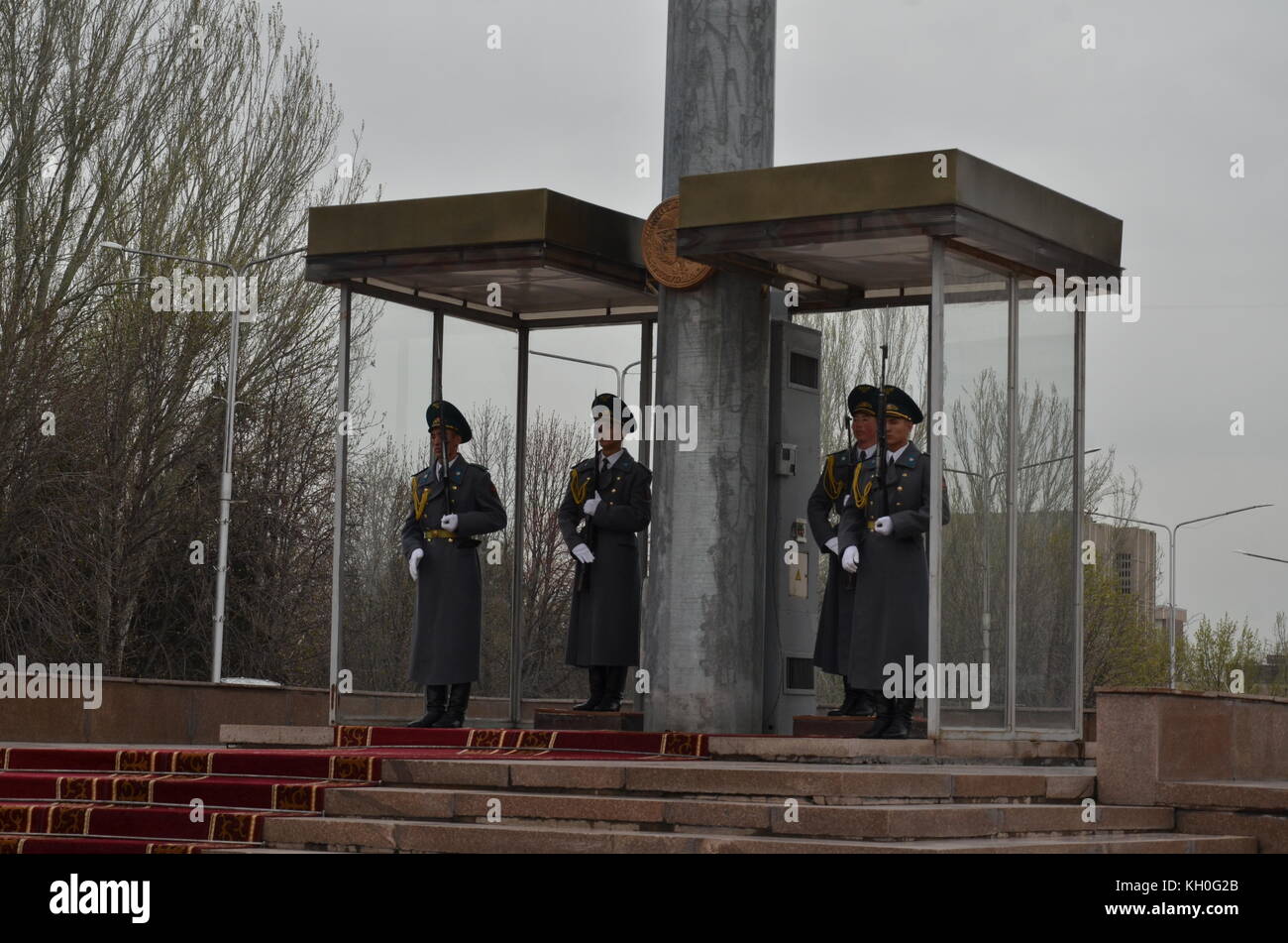 Cuatro guardias en uniforme con arma de pie junto a una enorme bandera polo con bandera nacional, Bishkek, Kirguistán, en Asia. Foto de stock