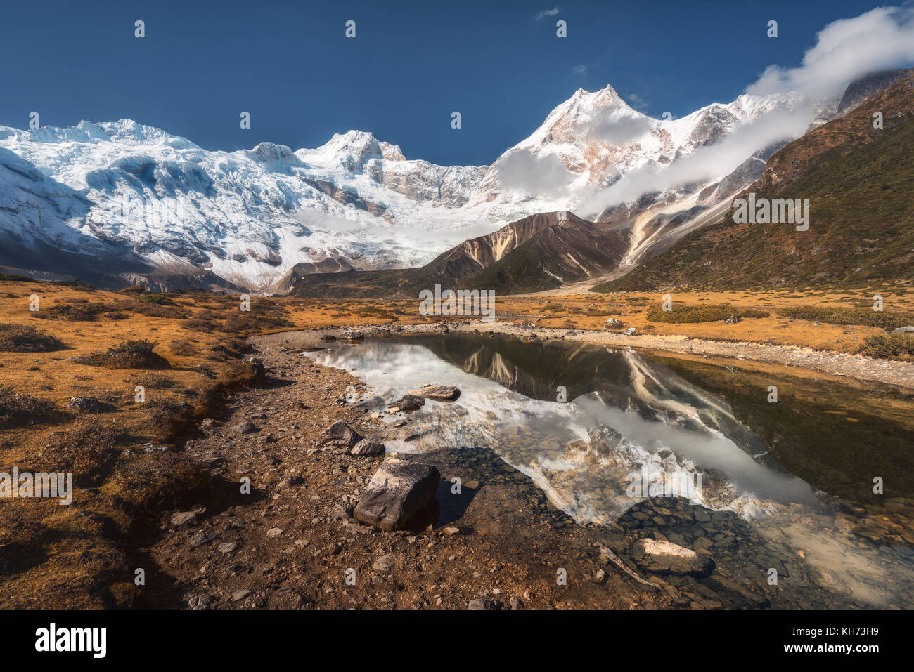 Hermosa vista con altas rocas con picos cubiertos de nieve, piedras en Mountain Lake, reflejo en el agua, el cielo azul con nubes en el amanecer. Nepal. Increíble Foto de stock