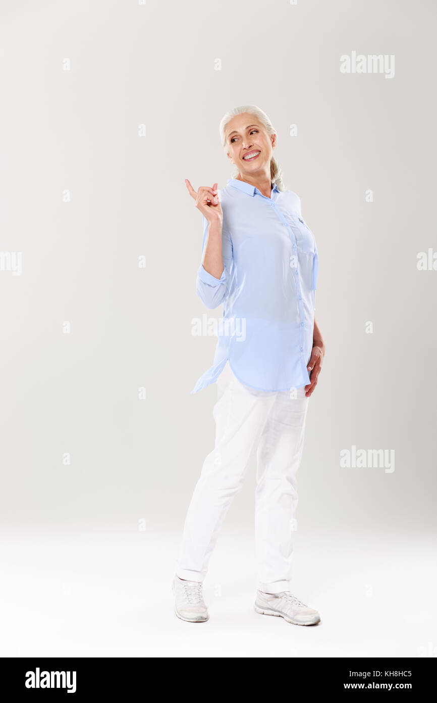Retrato de cuerpo entero de una mujer madura juguetona con camisa azul y pantalón  blanco, de pie y apuntando con el dedo, mirando a un lado, aislado en blanco  backgr Fotografía de