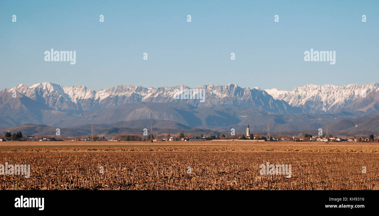 Un ex campo de maíz en diciembre en la región de Friuli, al noreste de Italia. Los Alpes Julianos y una iglesia de la aldea de spire son en el fondo Foto de stock