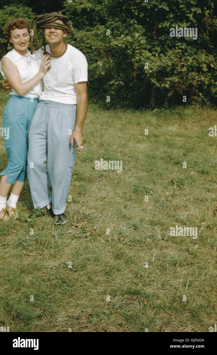 Una fotografía de un joven y una joven posando juntos, en un abrazo juguetón, El hombre tiene una gruesa cuerda enrollada alrededor de la parte superior de su cabeza y sus ojos, él es también el fumar un cigarrillo en la foto, están de pie en una zona ajardinada con árboles detrás de ellos, de 1952. Foto de stock