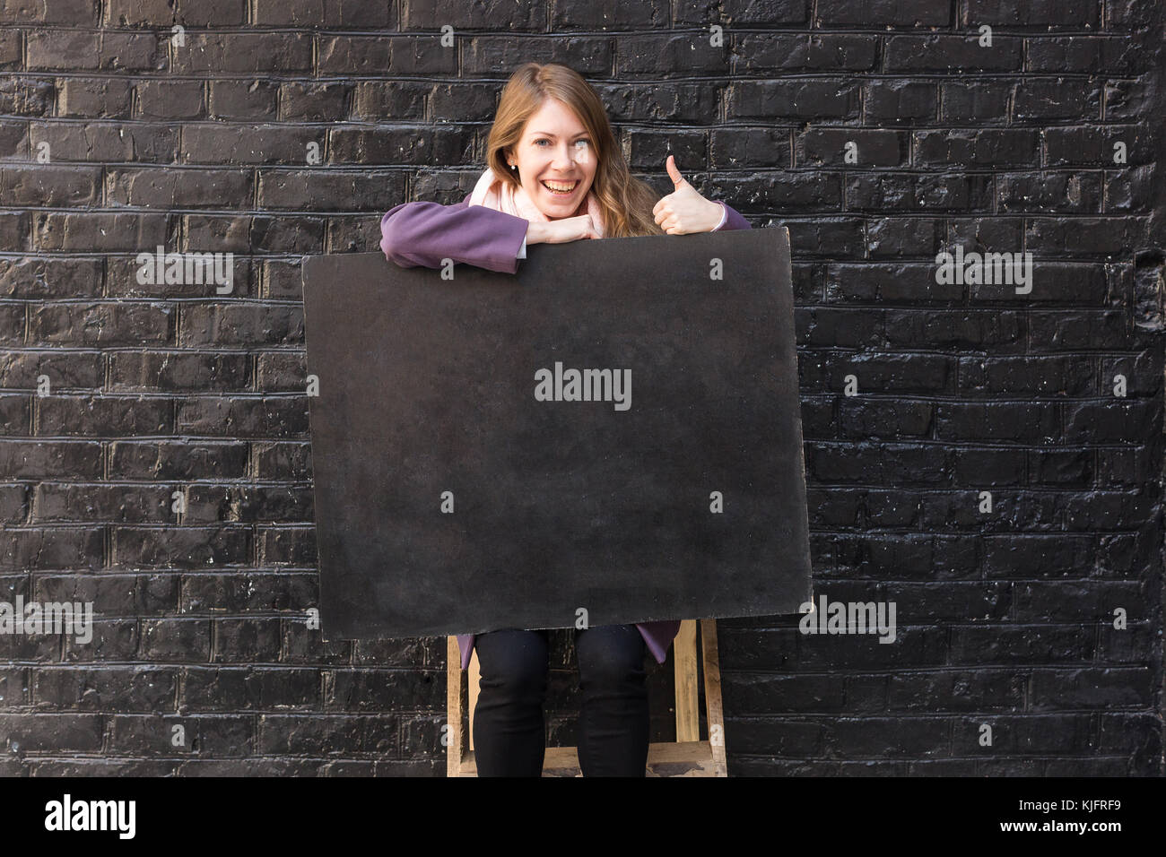 Antecedentes, artes y artesanías, emociones concepto. fairhaired joven loughing y dar pulgar arriba, ella tiene la sonrisa adorable y el anillo en el dedo pulgar, y blackboard sobre sus rodillas Foto de stock