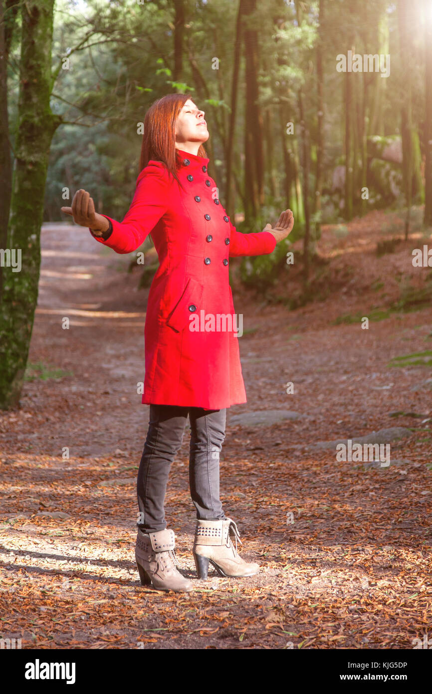 Mujer disfrutando de la calidez del sol de invierno solo en Forest Park ruta con brazos abiertos recibiendo rayos de luz, vistiendo un largo abrigo rojo o abrigo Foto de stock