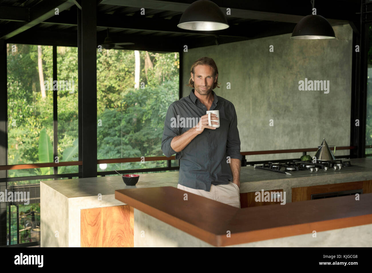 Guapo beber café en la cocina de diseño moderno con fachada de vidrio, rodeado por exuberantes jardines tropicales Foto de stock