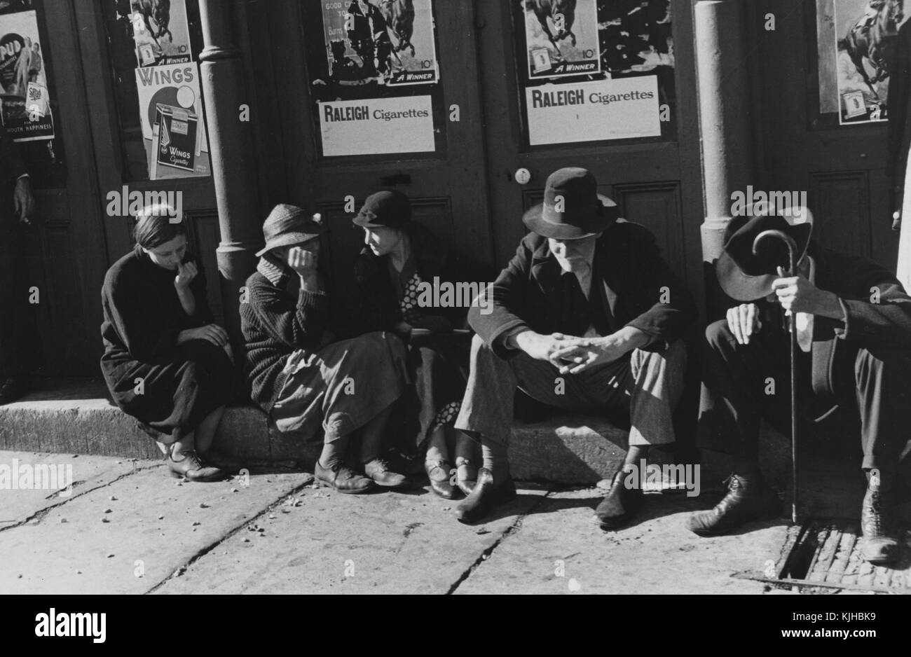 Una fotografía de tres mujeres y dos hombres sentados en una escalinata de concreto fuera de un edificio, los dos hombres llevan sombrero y vestimenta formal, las tres mujeres son todos luciendo vestidos y suéteres, estaban reunidos con otras personas como parte de una reunión religiosa, seis carteles que promocionan los cigarrillos están fijados a la parte exterior del edificio detrás de ellos, Tennessee, 1935. A partir de la biblioteca pública de Nueva York. Foto de stock