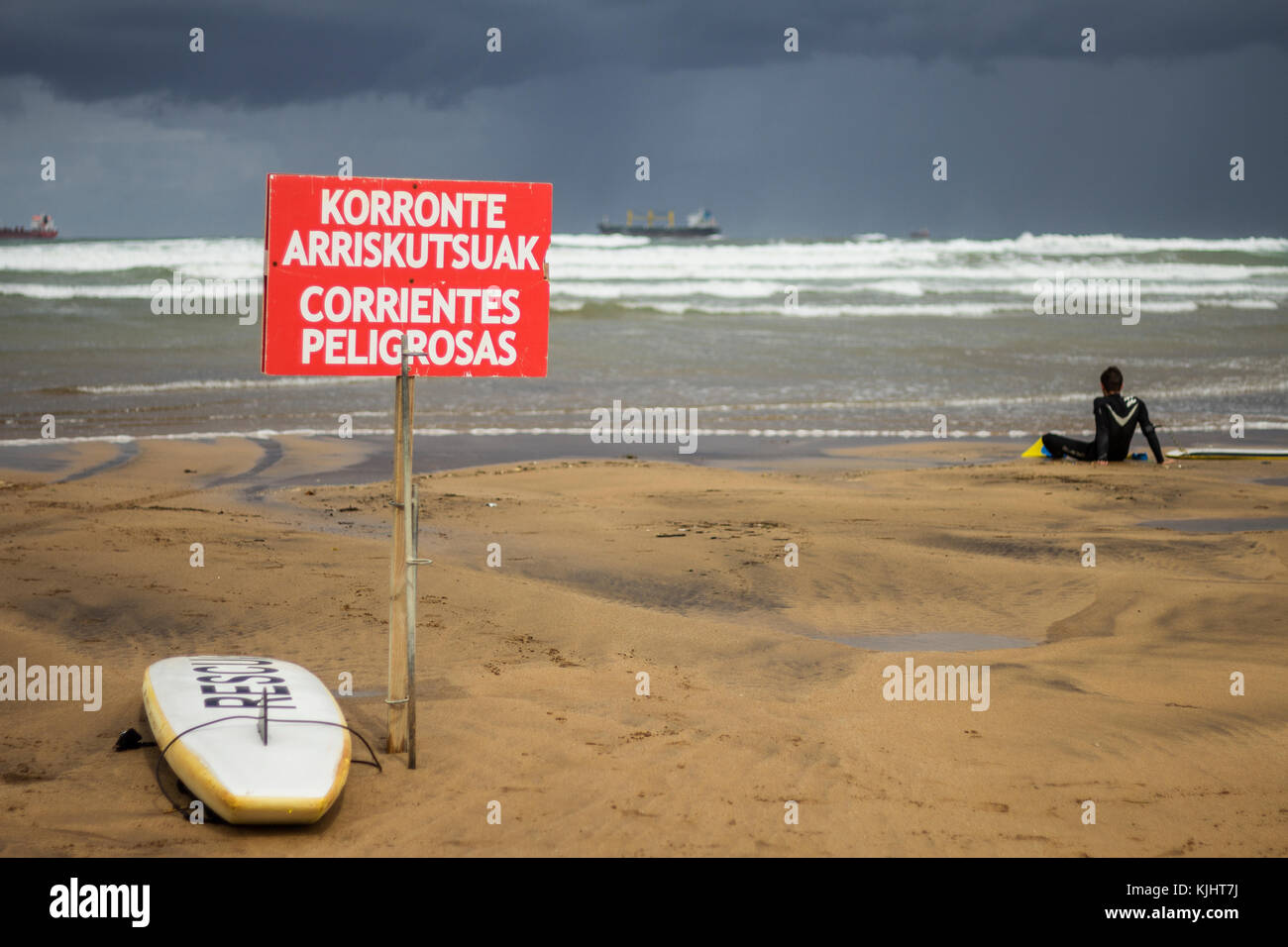 Surfista buscando la mala mar con una tabla de surf y una publicidad de corrientes peligrosas en una playa del país basue en españa Foto de stock