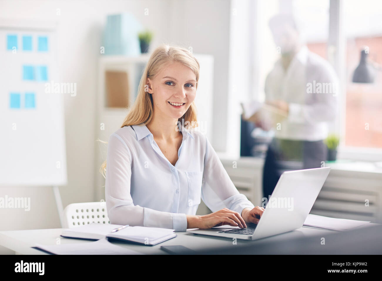 Secretario atractivo escribiendo en el teclado del portátil por el trabajo con el empresario de pie por la ventana al fondo Foto de stock