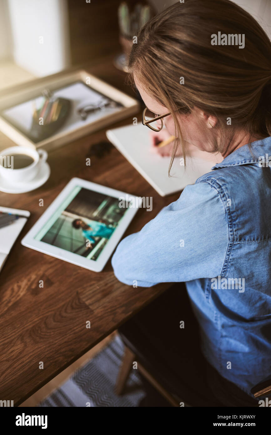 Mujer joven sentada sola en una mesa trabajando con una tableta digital y escribir las ideas en un bloc de notas Foto de stock