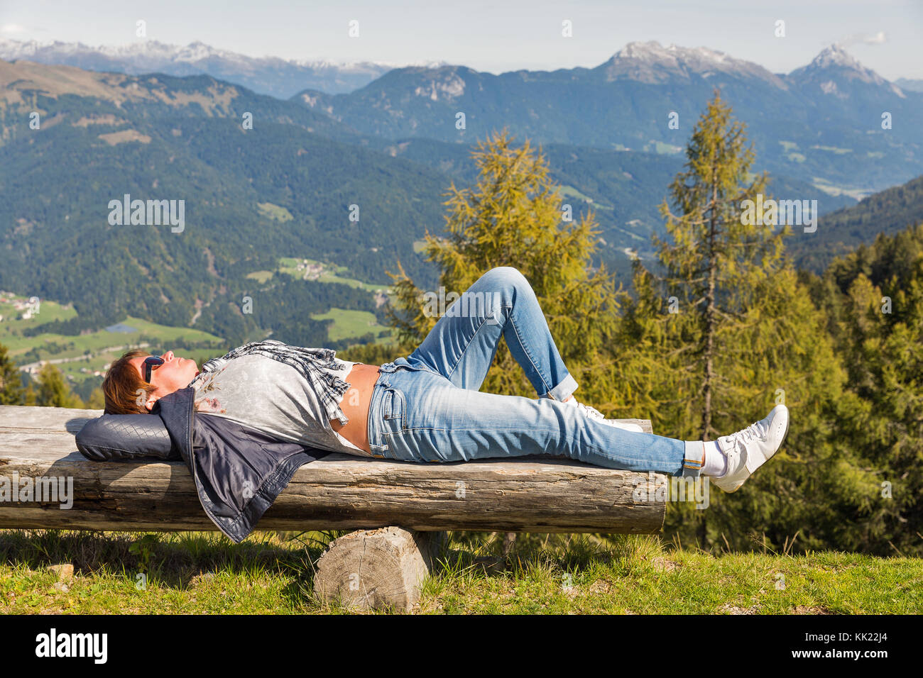 Mujer de mediana edad caucásico tomar el sol y descansar con las manos detrás de la cabeza en el banco de madera con vistas al paisaje de las montañas alpinas en el oeste carint Foto de stock