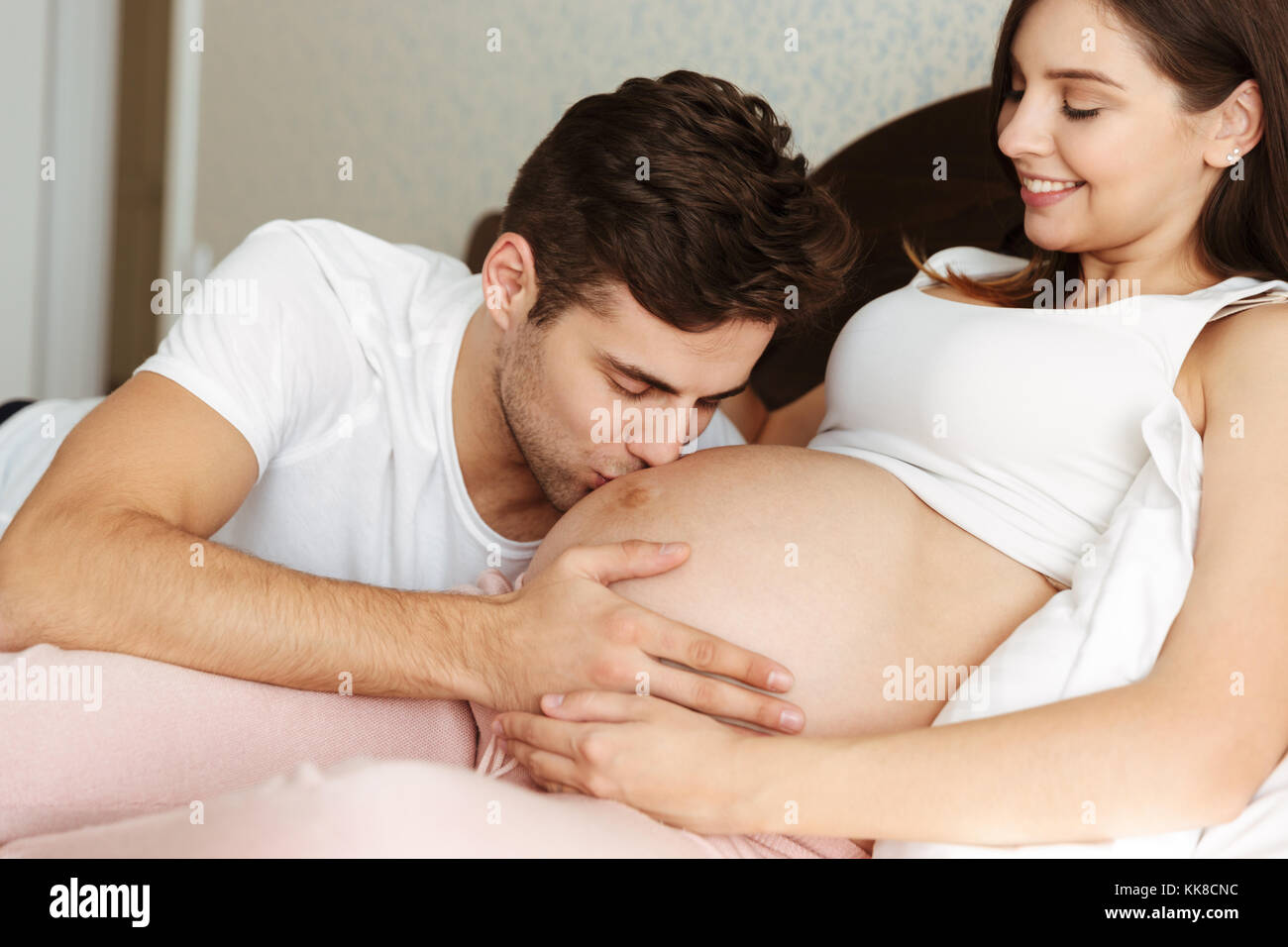 Sonriente joven esposa embarazada acostado en la cama con su marido en el dormitorio, el hombre besa su vientre Foto de stock