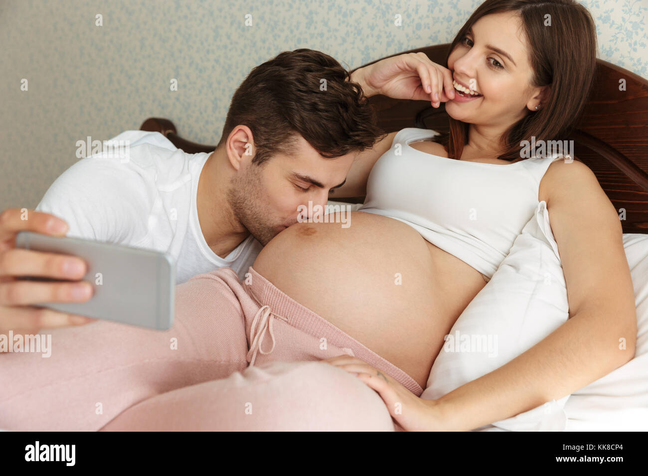 Adorable joven pareja embarazada tomando selfie tumbado en la cama, el hombre bese embarazada vientre Foto de stock