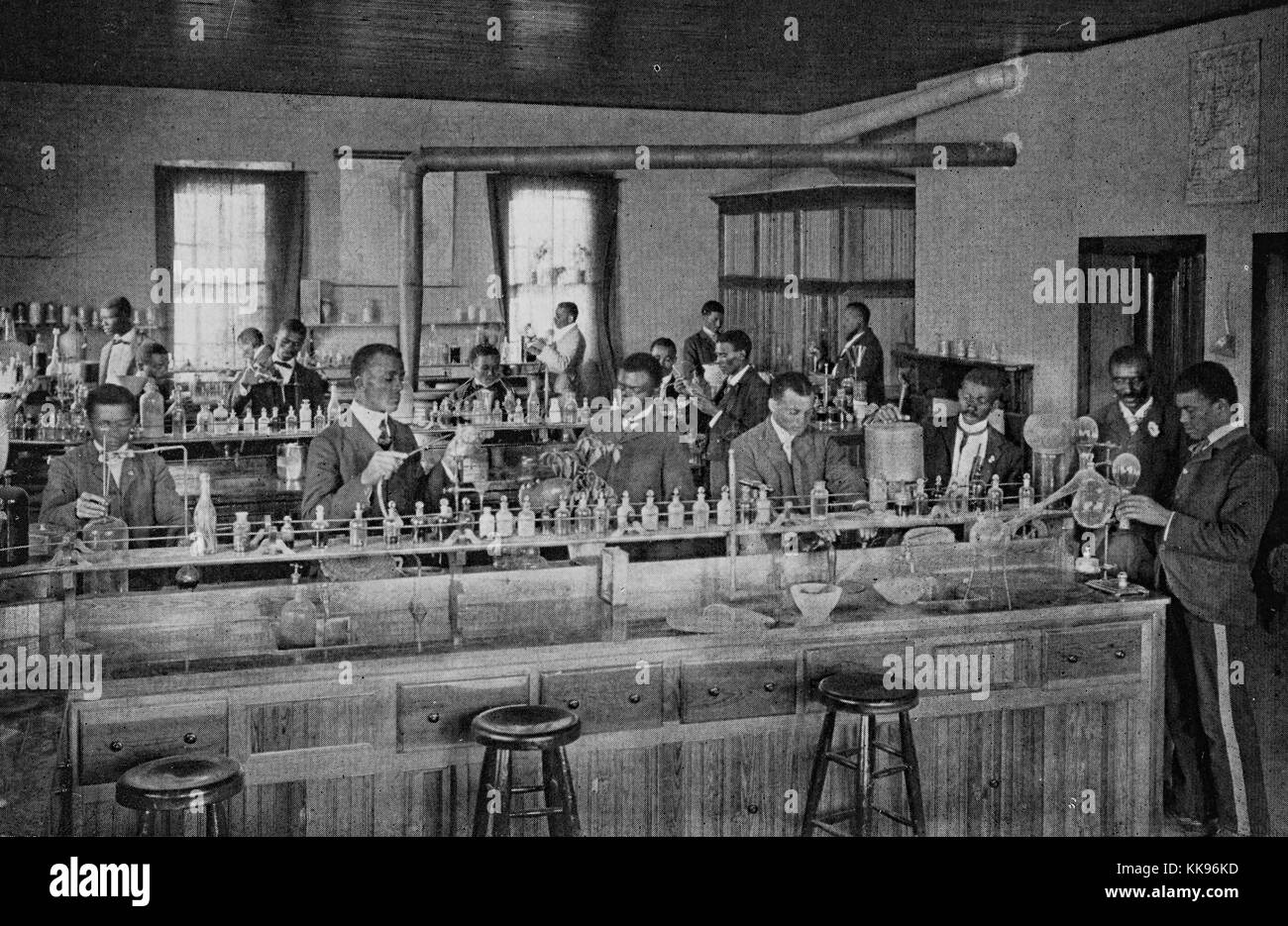 Fotografía en blanco y negro de un grupo de hombres afroamericanos, todos  en trajes, trabajando en largas mesas con botellas de reactivos y equipo de  vidrio, en un laboratorio agrícola, 1904. Desde