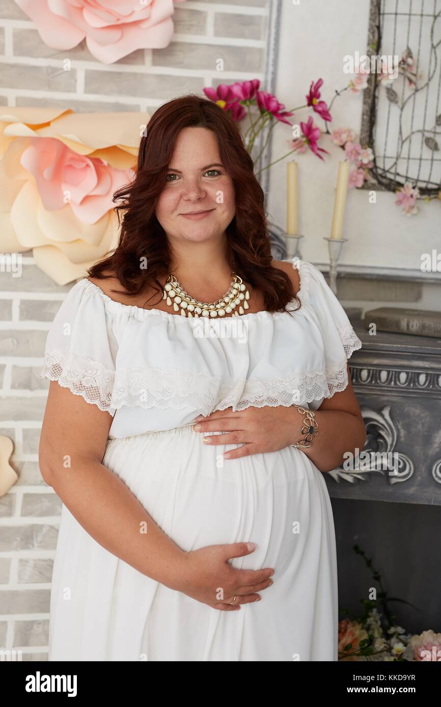 Adorable mujer embarazada con alegre sonrisa en vestido blanco puro y joyas  abraza su vientre alojarse en decoración interior Fotografía de stock -  Alamy
