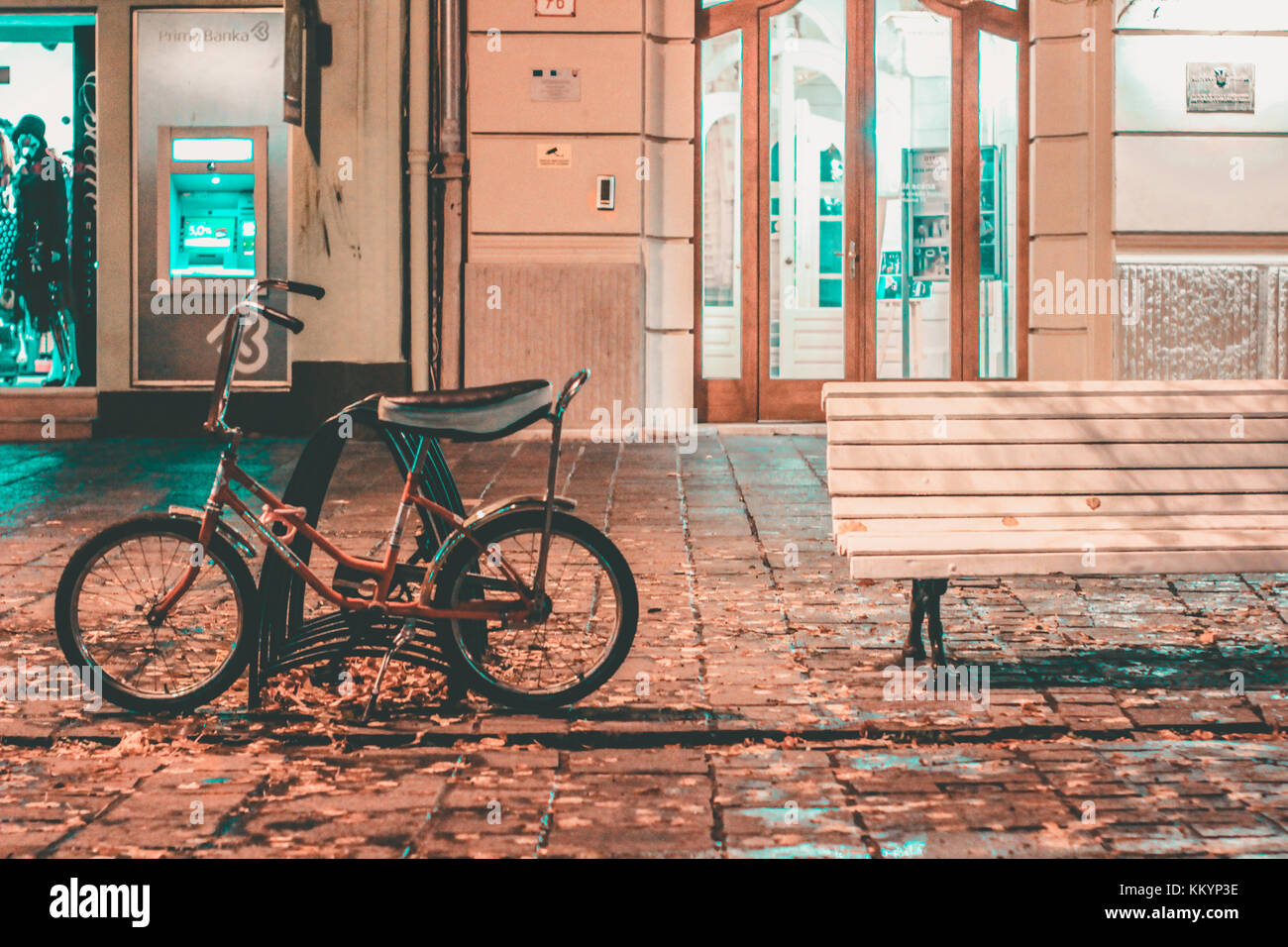 Bicicleta cerca de un pupitre vacío fuera un cajero y tiendas en una carretera asfaltada paisaje nocturno el verde azulado y naranja Foto de stock