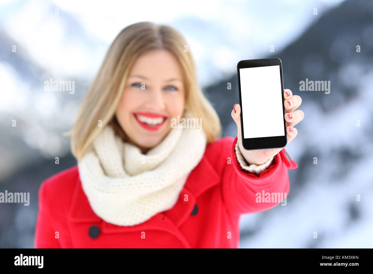 Vista frontal retrato de una niña en rojo muestra un espacio en blanco de la pantalla del teléfono inteligente en una montaña nevada en invierno Foto de stock