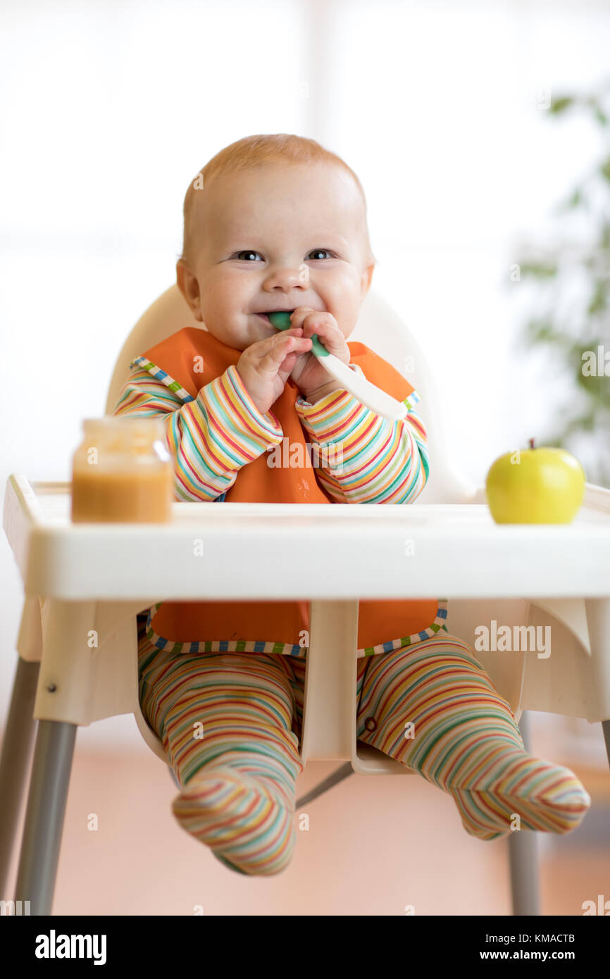 Alegre bebé come alimentos con cuchara. Retrato de niño feliz muchacho en silla alta. Foto de stock