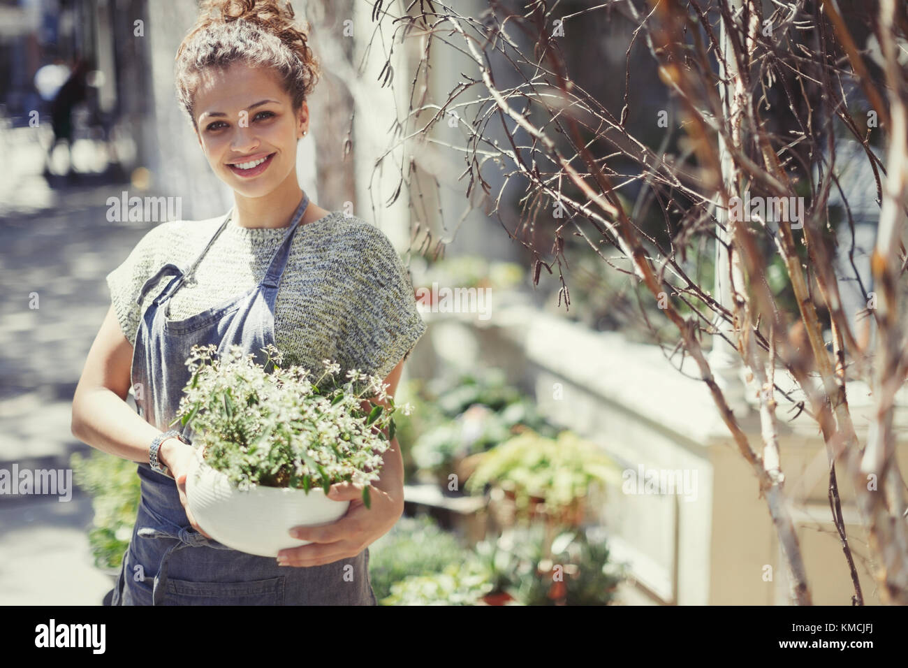 Retrato sonriente floristería femenina sosteniendo planta en macetas en el soleado escaparate Foto de stock