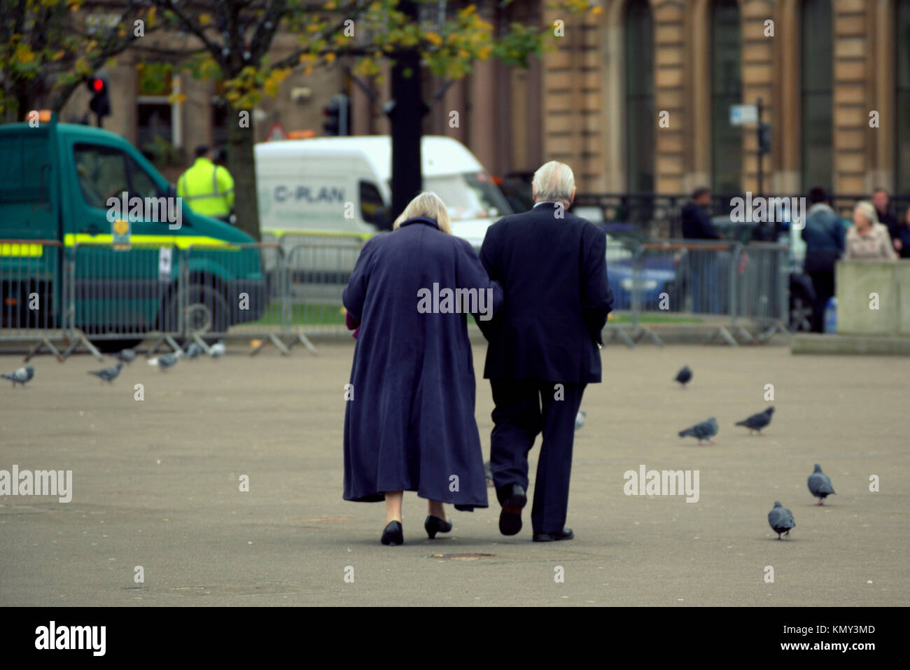 Pareja de ancianos ancianos hombre y mujer mujer vistiendo el azul marino a través de la plaza caminando entre las palomas visto desde detrás de George Square, Glasgow, Reino Unido Foto de stock