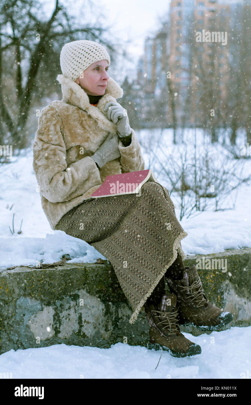 Una mujer adulta con un abrigo de piel y un gorro de lana está sentado en  una banca de piedra y leer un libro en invierno, en un parque cubierto de  nieve