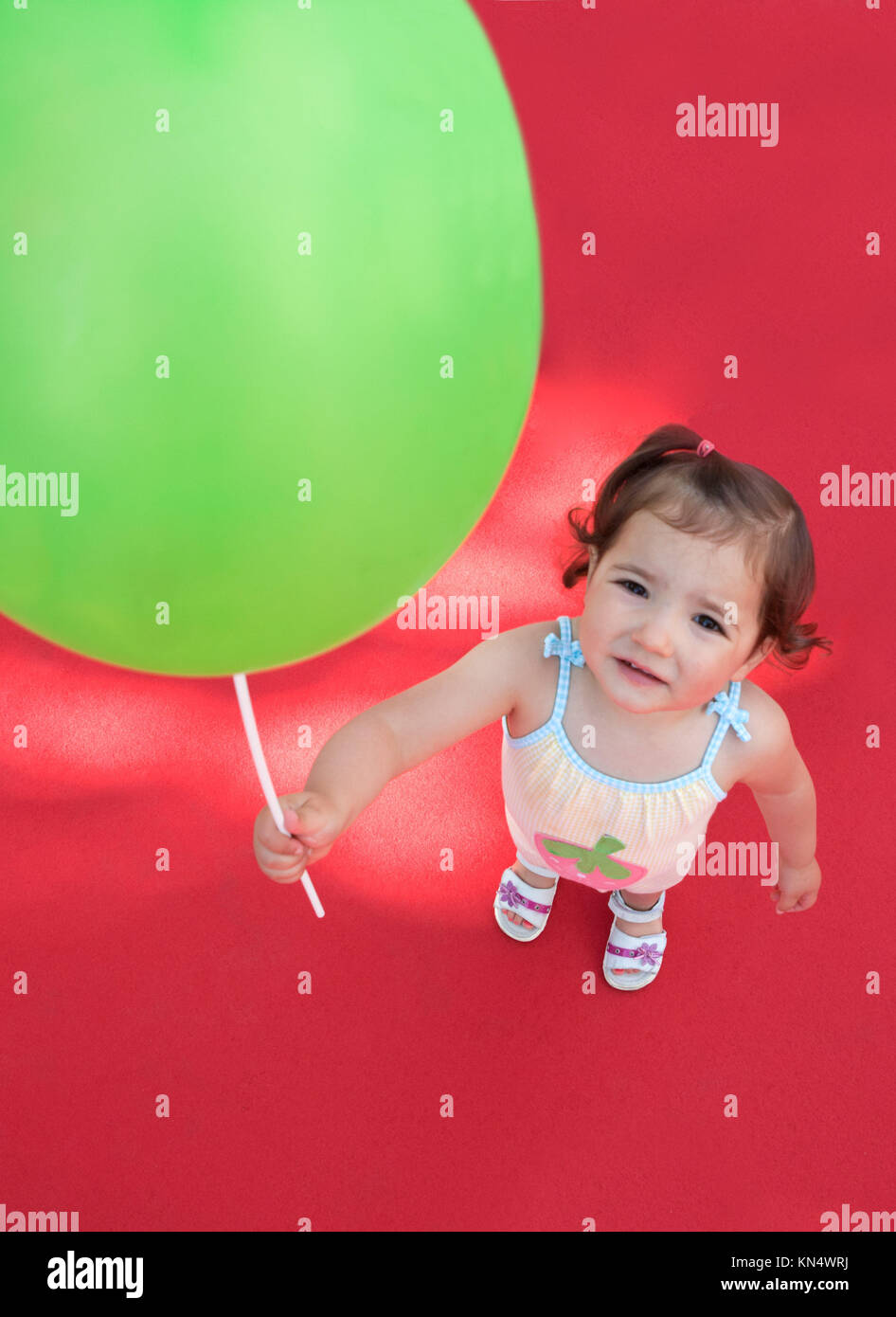 Una niña afuera sosteniendo un globo verde sobre una alfombra roja. Un alto ángulo de disparo. Foto de stock