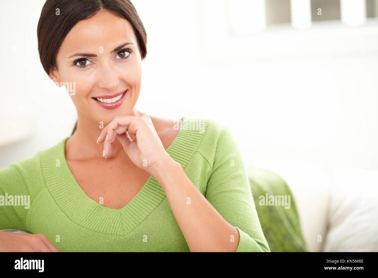 Sonriente mujer caucásica en camisa verde mirando a la cámara mientras se pone la mano en la barbilla en interiores - copia el espacio. Foto de stock