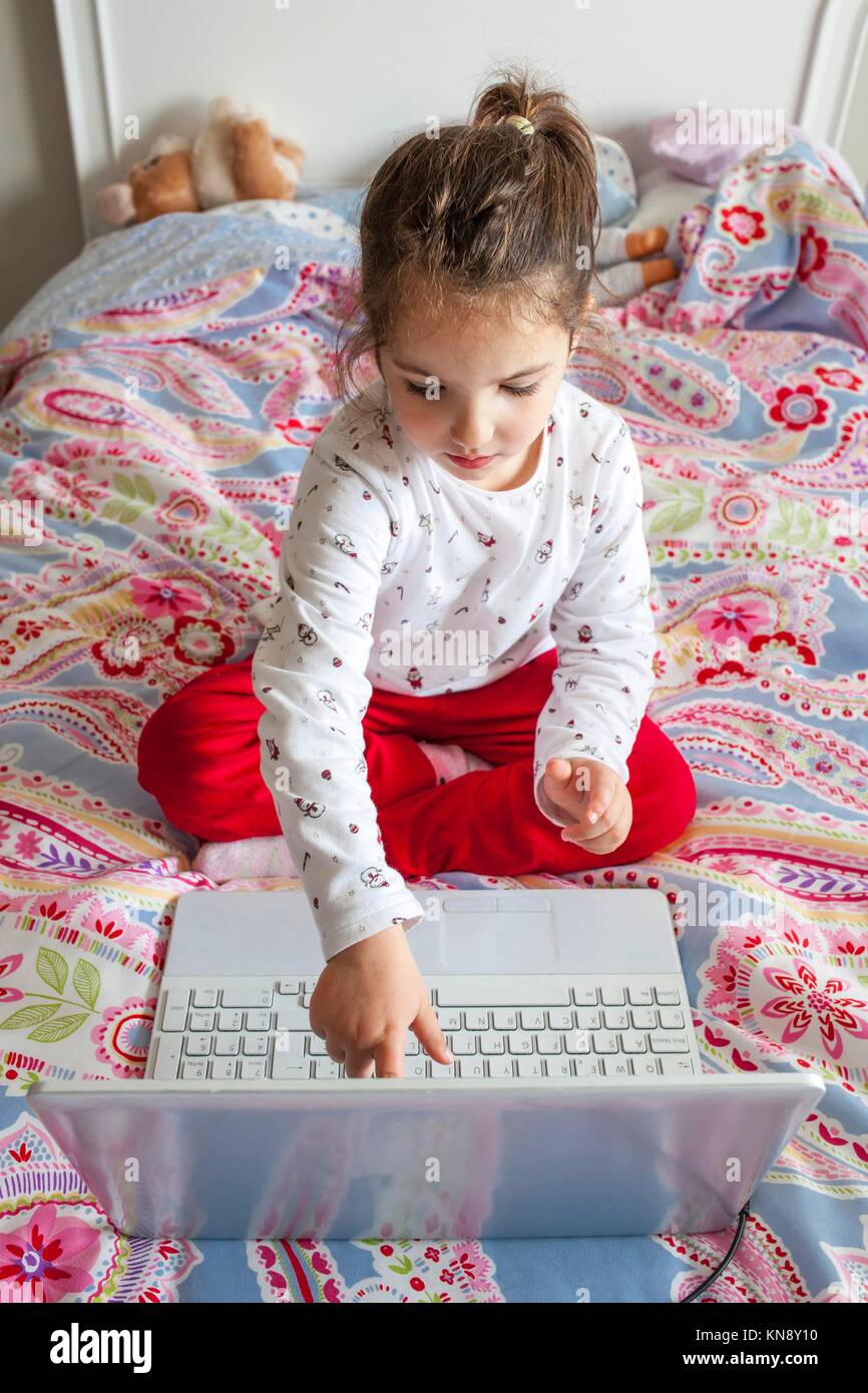 Niña sentada en la cama y jugar en línea con el ordenador portátil. Ella está tocando la pantalla. Foto de stock