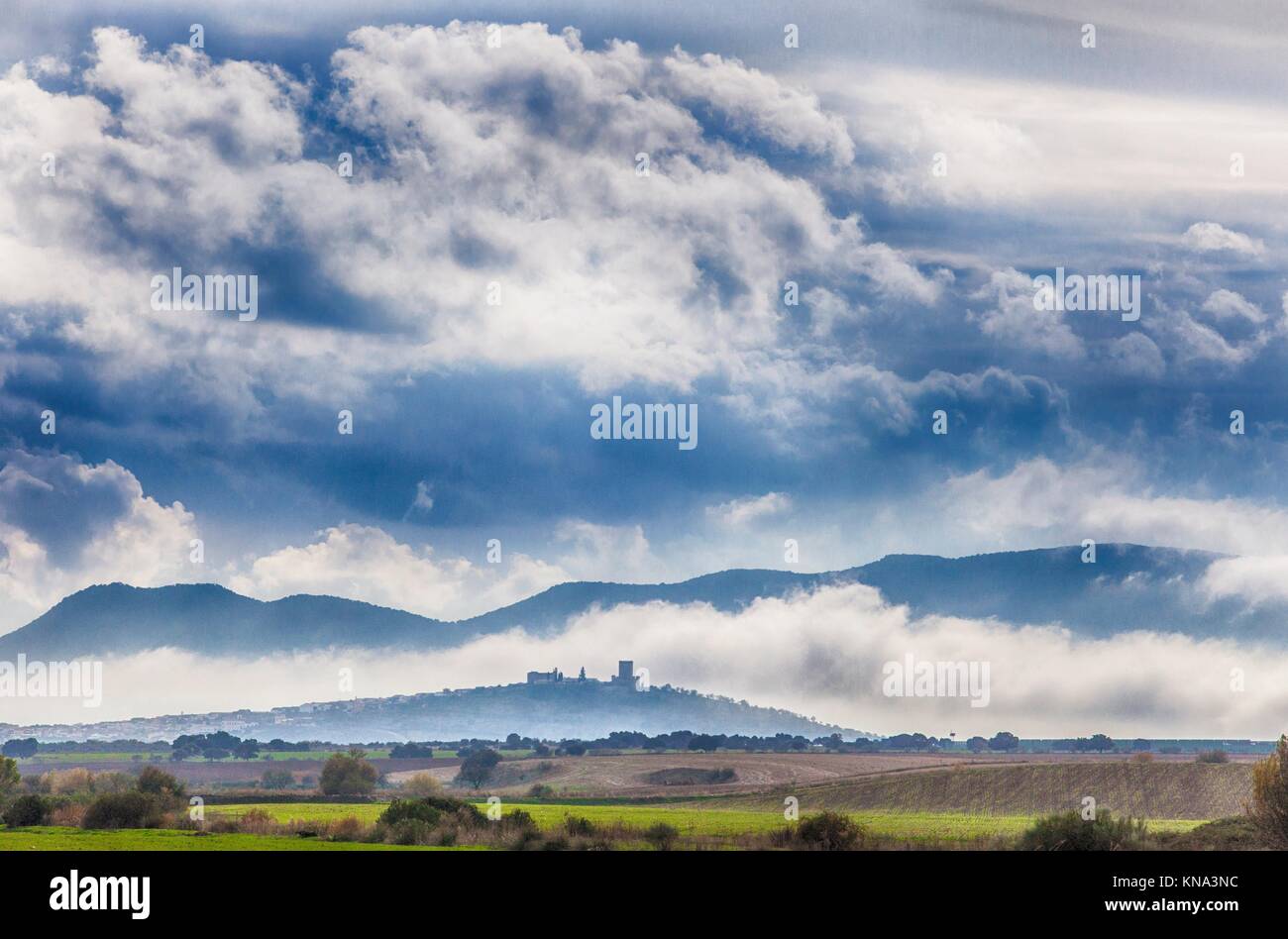 A primera hora de la tarde el paisaje de campos de cereales y pequeña ciudad con castillo, entre la niebla, Nogales, España. Foto de stock