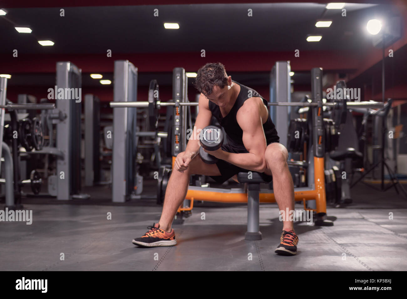 Un hombre joven, gimnasio oscuro en interiores, equipos de gimnasia, una mano pesa bíceps ejercicio, banco para sentarse. Foto de stock
