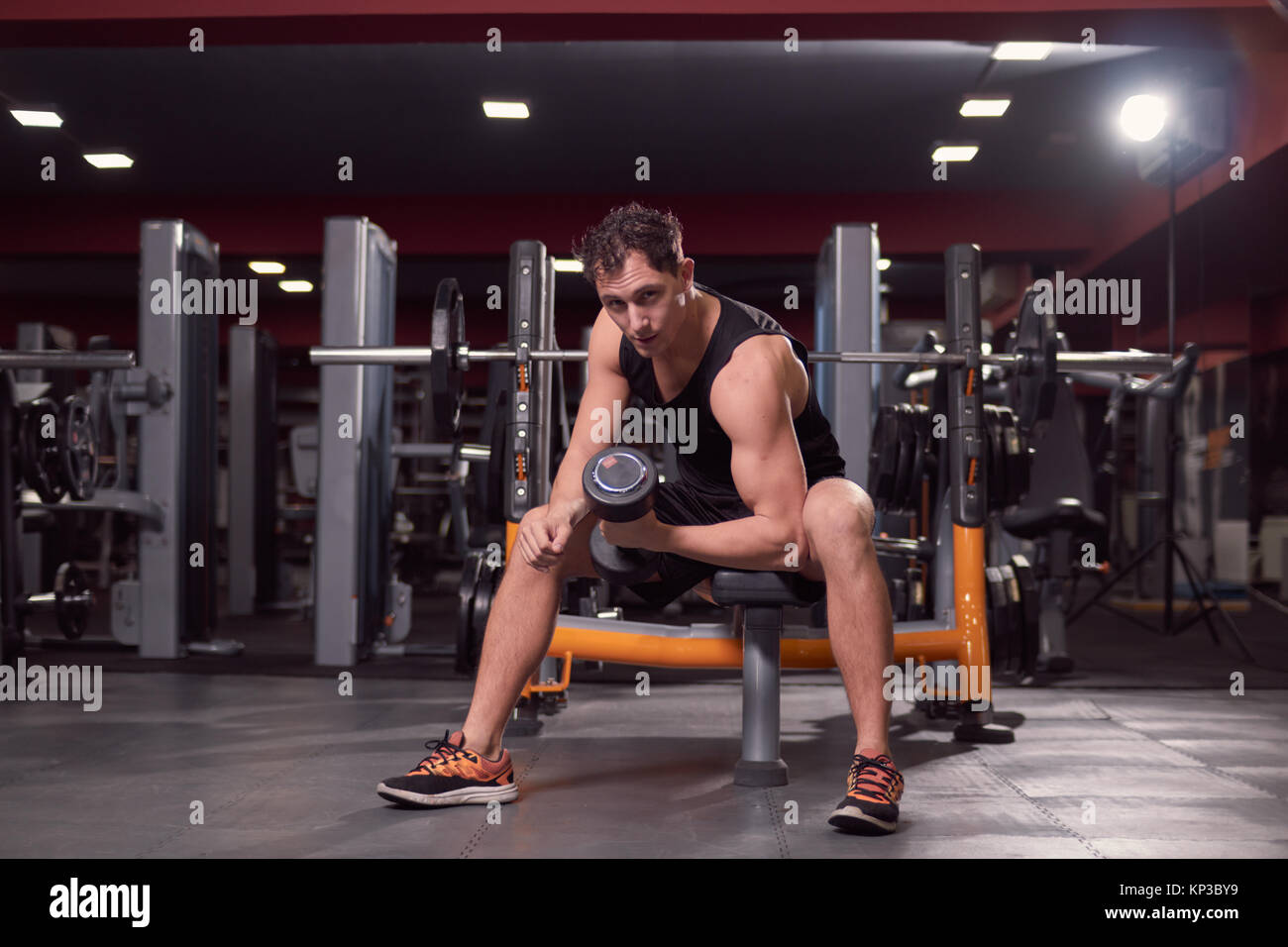 Un hombre joven, gimnasio oscuro en interiores, equipos de gimnasia, una mano pesa bíceps ejercicio, banco para sentarse, mirando a la cámara. Foto de stock