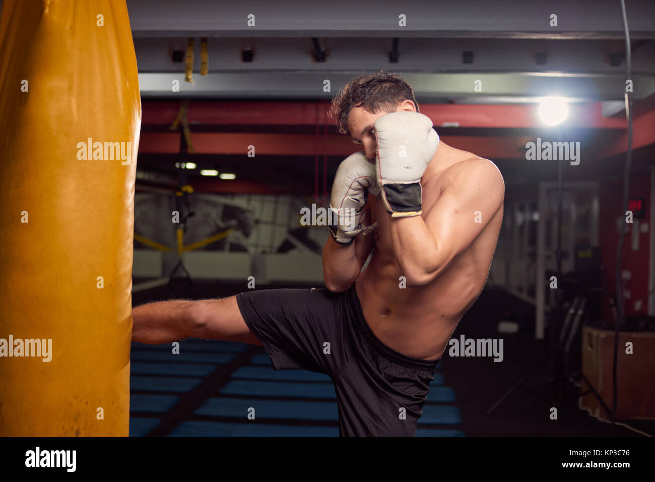 Un joven hombre descamisado, boxeador pierna golpeando saco de boxeo, practicando en interiores, sala de gimnasia, usando guantes de boxeo, disparos en la parte superior del cuerpo. Foto de stock